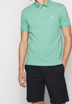 Zielona bawełniana koszulka polo z krótkimi rękawami, kołnierzykiem na guziki i małym haftowanym logo, zestawiona z ciemnoniebieskimi szortami.