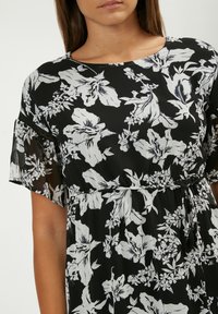 Schwarzes Kleid mit weißem Blumenmuster, kurzen semi-transparenten Ärmeln, runder Halslinie und Taillengurt. Weicher, fließender Stoff.
