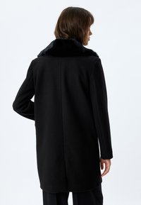 Cappotto di lana nera con colletto in pelliccia sintetica, taglio dritto, maniche lunghe e orlo pulito. Nessuna tasca o bottone visibile.