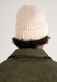 Person with curly dark hair wearing a cream-colored ribbed knit beanie and an olive green leather jacket, viewed from behind.