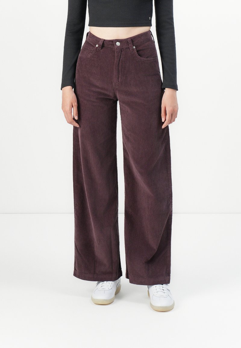 Persoon in donkerpaarse corduroypantalon met hoge taille en wijde pijpen, gedragen met witte sneakers en een zwart langemouwen crop-top tegen een witte achtergrond.