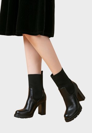Bottes noires en cuir jusqu'à la cheville avec un dessus en chaussette côtelée noir, un talon bloc et une semelle texturée. Fini brillant avec un design épuré.