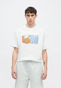 Λευκό βαμβακερό μπλουζάκι με πολύχρωμο γραφικό που απεικονίζει ένα μπάσκετ και έναν χαρακτήρα κινουμένων σχεδίων. Κοντά μανίκια, άνετη γραμμή, συνδυασμένο με γκρι σορτς.