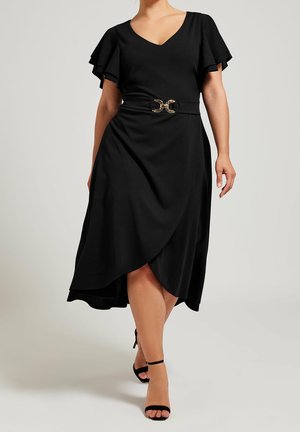 Woman walking wearing a black V-neck dress with flutter sleeves, wrap skirt, gold belt buckle, and black ankle-strap heels.