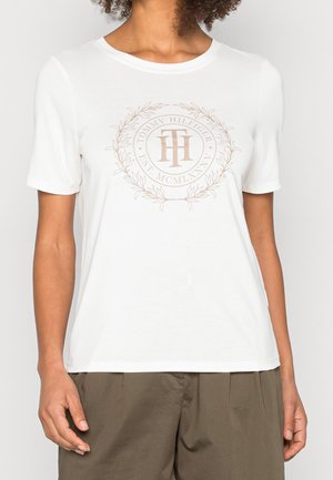 Kobieta w białej koszulce z krótkim rękawem z beżowym logo Tommy Hilfiger i oliwkowo-zielonych spodniach.