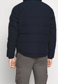 Granatowa pikowana kurtka z kołnierzem na zamek, z przeszywanymi sekcjami i naszywką z logo na lewym ramieniu. Nosi się ją z szarymi spodniami bojówkami.