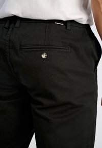 Primer plano de una persona que lleva pantalones negros con un bolsillo trasero abotonado y una camisa blanca metida por dentro.