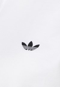 Czarne logo trefoil Adidas wyśrodkowane na gładkim, białym, teksturowanym tle tkaniny.