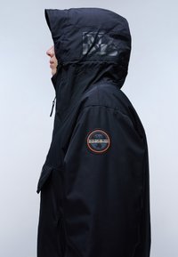 Chaqueta negra aislante con un alto cuello, capucha ajustable, bolsillos con cremallera y un parche redondo con logo en la manga izquierda. Textura suave.