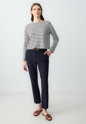 Chemise à manches longues rayée en crème et bleu marine, associée à un pantalon bleu marine ajusté. Une ceinture marron et des sandales tissées marron complètent la tenue.