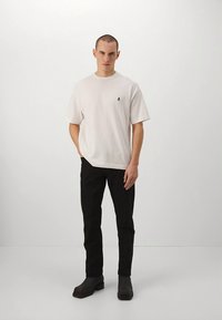 Pánské bílé tričko s krátkým rukávem a malým černým logem na hrudi, doplněné černými kalhotami a černými kotníkovými botami.