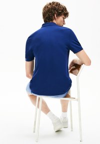 Donkerblauwe polo met korte mouwen, voorzien van een rechte zoom en kraag, gedragen met lichte denim shorts en witte sneakers terwijl zittend.