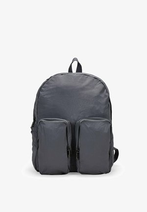 Mochila de tela gris con forma redondeada, dos bolsillos frontales con cremallera y un asa en la parte superior. Superficie ligeramente texturizada. Correas ajustables.