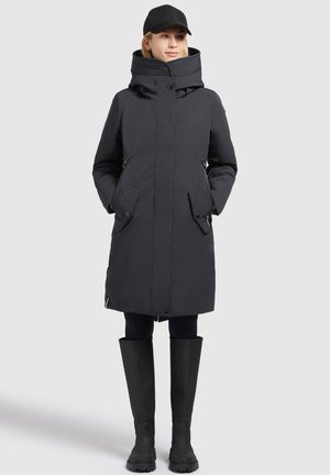 Parka nera trapuntata con colletto alto, tasche laterali e chiusura con zip. Abbinata a stivali di gomma neri alti. Design semplice e funzionale.