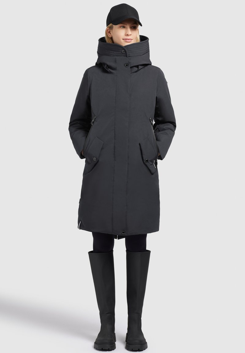 Parka nera con colletto alto, bottoni nascosti e tasche con zip. Abbinata a leggings aderenti e stivali di gomma neri alti.