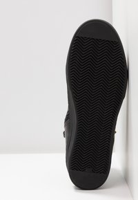 Semelle de chaussure noire avec motif de bande en chevron reposant verticalement contre un coin blanc de mur et de sol.