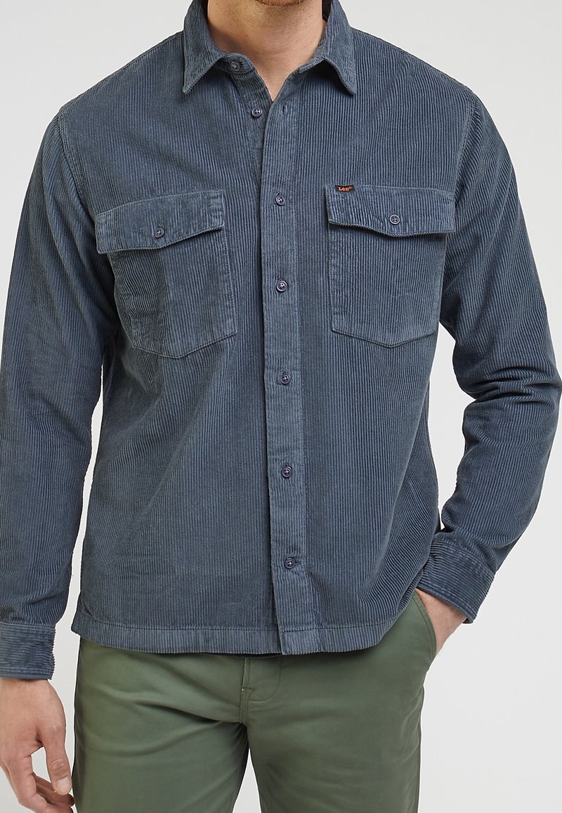 Camicetta in corduroy blu scuro con due tasche sul petto, chiusura a bottoni e un motivo a righe testurizzato. Vestibilità slim e design del colletto classico.