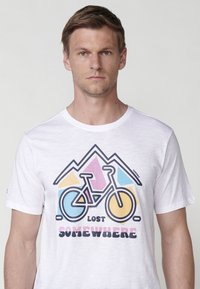 White cotton t-shirt featuring a bicycle graphic. Design includes mountains and pastel accents—pink, yellow, and blue. Text: "LOST SOMEWHERE."