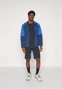 Veste coupe-vent bleue et navy avec capuche ajustable, zips et poignets élastiques, portée sur un t-shirt noir et un short sombre.