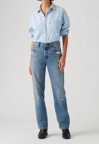 Person som bär en ljusblå skjorta med uppkavlade ärmar, högmidjade slitna blå jeans och svarta ankelboots mot en enfärgad bakgrund.