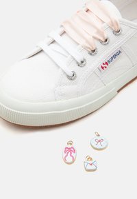 Baskets blanches Superga avec de larges lacets en satin et trois petits charms en dessous représentant un nœud rose, un nœud bleu et des motifs d’enveloppe.