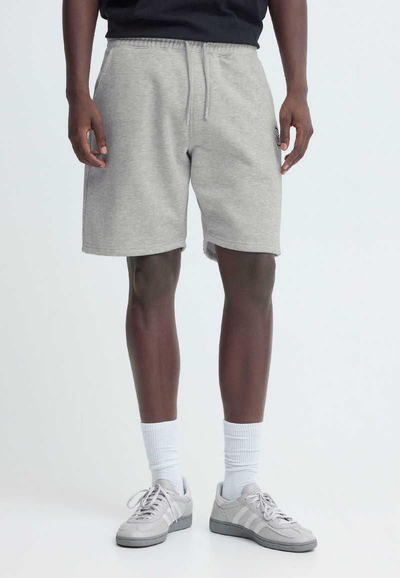 Shorts en mélange de coton gris avec une taille élastique et un cordon de serrage. Dotés de poches latérales et d'un logo discret. Portés avec des baskets grises et des chaussettes blanches.