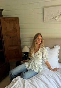 Femme blonde en blouse fleurie et jean, assise sur un lit avec du linge de lit blanc, à côté d'une armoire en bois et d'une lampe de chevet.