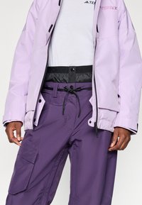 Chaqueta impermeable de color púrpura claro con detalles en negro sobre una camiseta blanca, combinada con pantalones cargo morados que cuentan con cintura ajustable y bolsillos.
