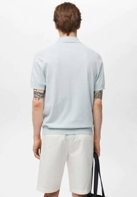 Tricou polo cu mânecă scurtă de culoare albastru deschis afișat din spate, având o textură netedă și un tiv ribotat. Pantaloni scurți albi și o geantă închisă la culoare sunt vizibile.
