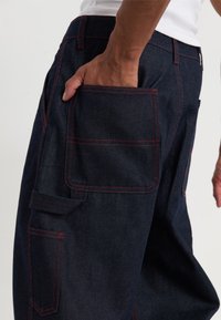 Pantalones de mezclilla en azul oscuro con costuras rojas, que cuentan con múltiples bolsillos, incluido un gran bolsillo lateral, diseñados para la utilidad y la funcionalidad.