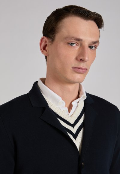 Navy knit blazer with a notched lapel, worn over a white shirt and a cream sweater featuring navy stripes. Smooth texture and tailored fit.