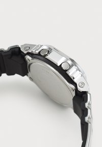 Montre-bracelet en acier inoxydable argenté avec un bracelet en caoutchouc noir. Comprend deux boutons sur les côtés et un matériel visible. Dos lisse avec un design circulaire.