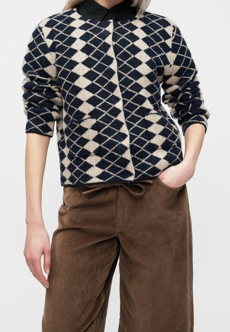 Personne portant un pull à motif losanges noir et beige sur une chemise noire à col, un pantalon en velours côtelé marron, une main dans la poche.