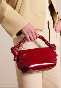 Red patent leather handbag with a knotted handle. The bag has a rounded shape and a glossy finish, accentuating its smooth texture.