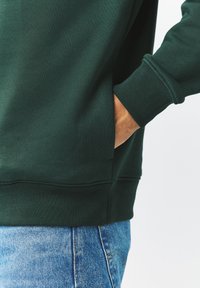 Tmavě zelená mikina s kulatým výstřihem, žebrovanými rukávy a bočními kapsami. Doplněná světle modrými riflovými kalhotami. Hladká textura.