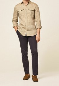 Chemise beige en lin à boutons avec deux poches poitrine, associée à un pantalon sombre et des mocassins marron. La chemise a des manches retroussées et une coupe décontractée.