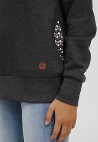 Siva pot sweatshirt z žepom s cvetličnim vzorcem in rjavim logotipom. Ima rebraste manšete in sproščen kroj. Nosi se čez modre traperice.