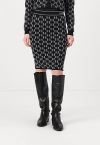 Jupe crayon en tricot noire et gris avec un motif de logo répétitif, associée à des bottes noires en cuir jusqu'aux genoux.