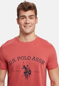 Crvena pamučna majica s okruglim izrezom, kratkim rukavima i središnjim grafičkim prikazom igrača poloa uz tekst "U.S. POLO ASSN." u mornarsko plavoj boji.