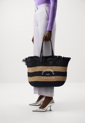 Osoba w lawendowych spodniach i fioletowej bluzce z długim rękawem trzyma czarną i beżową torbę plecioną z napisem, widoczne srebrne buty na wysokim obcasie.