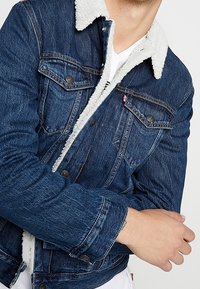 Jeansjacka i mörkblå färg med fårskinnskrage, knäppning framtill, påsydda fickor och kontrasterande sömmar längs med sömmarna.