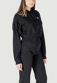 Felpa nera corta con tasca frontale e cappuccio con cordino, abbinata a pantaloni della tuta neri coordinati con vita elastica.