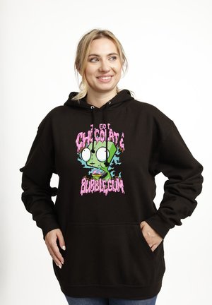 Femme souriante portant un sweat à capuche noir avec un personnage de dessin animé vert et un texte rose "Chocolate Bubblegum" sur le devant, les mains dans les poches.