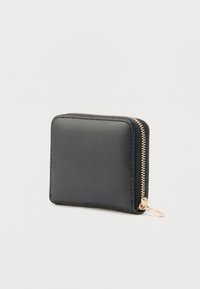 Cartera compacta de cuero negro con cierre de cremallera dorada, mostrada cerrada sobre un fondo claro y simple.