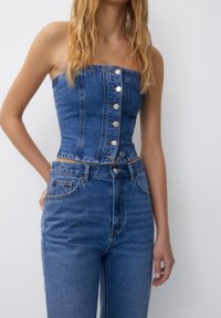 Top de bustier de mezclilla con corte estructurado, que presenta un escote recto, siete botones frontales y color azul claro, combinado con jeans de cintura alta.
