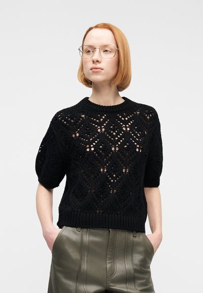 Tânăr cu păr scurt roșu și ochelari, purtând un pulover negru tricotat cu model și pantaloni din piele verde măsliniu, cu mâinile în buzunare.