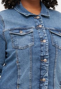 Veste en denim bleu clair avec poches poitrine, boutons en métal argenté et surpiqûres contrastantes. Dispose d'un col classique et d'une coupe décontractée.