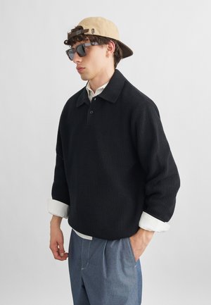 Junger Mann mit einer beigefarbenen Rückwärtskappe, dunkler Sonnenbrille, schwarzem Strickpullover über einem weißen Hemd und blauer Hose, eine Hand in der Tasche.