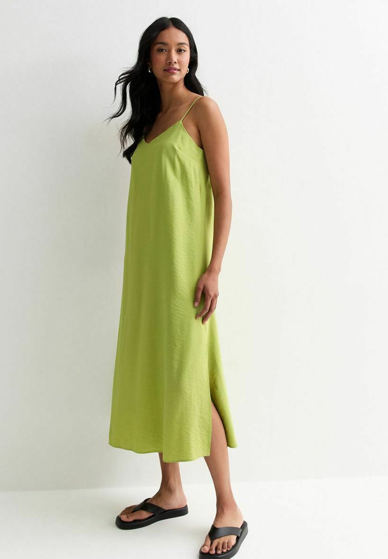 Groene, mouwloze maxi-jurk met een V-hals, een zijsplit en een zachte textuur. Gecombineerd met zwarte teenslippers tegen een lichte achtergrond.