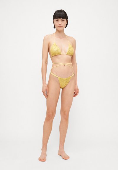 Bikini amarillo con lentejuelas, con un top triangular y cobertura mínima, presentando tirantes delgados que se enroscan alrededor de la cintura y las caderas.
